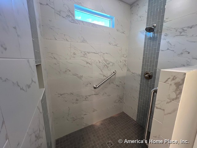 Walk-in shower with marble-look wall tile, vertical accent strip, built-in grab bar, gray tile floor, recessed niche shelf, and transom window above.