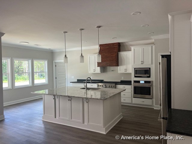 White shaker cabinets, granite island countertop with built-in stainless oven and microwave, wood range hood, pendant lighting fixtures, wide plank flooring, large windows, and