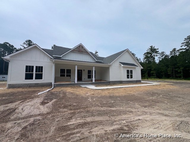 Covered front porch with white siding exterior, multiple large windows, gable roof, decorative trim details, entry door, and unfinished landscaping in new residential construction.