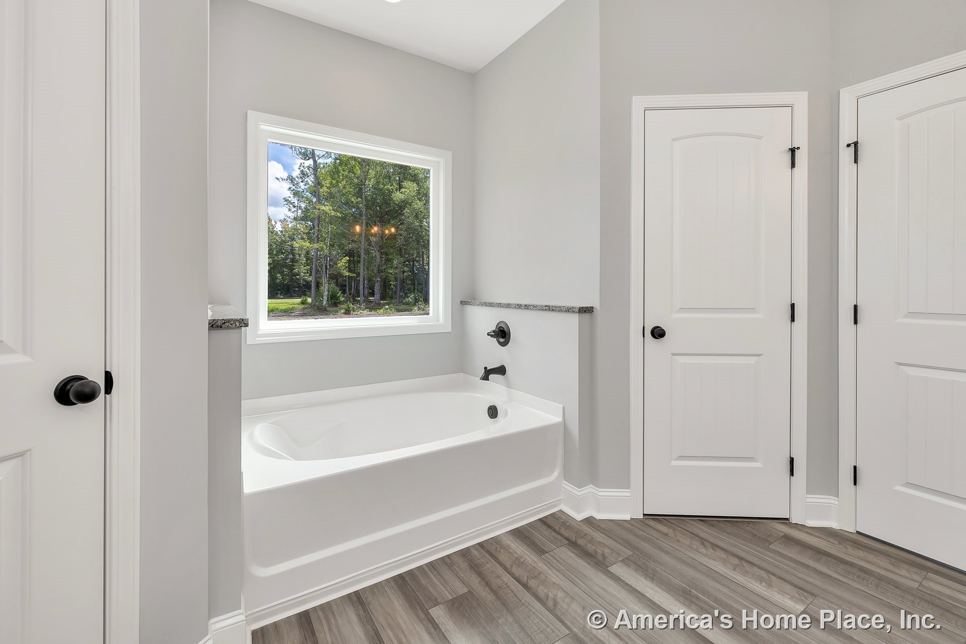 Built-in soaking tub with black fixtures set beneath a large picture window, wood-look tile flooring, white paneled doors, granite ledge trim, light gray walls, and baseboard trim.