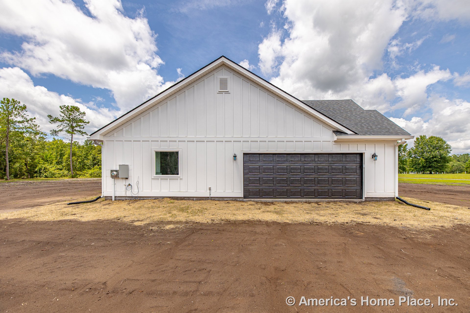 White vertical board and batten siding with dark paneled two-car garage door, single rectangular window, gable roof with asphalt shingles, exterior wall-mounted lanterns, and vent