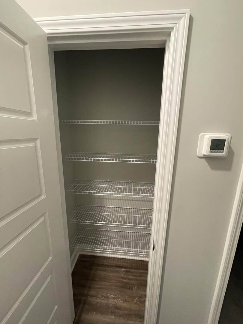 Walk-in closet with white shelves, wooden floor, white walls and baseboard, door with silver handle, and a small white device with screen mounted on the wall