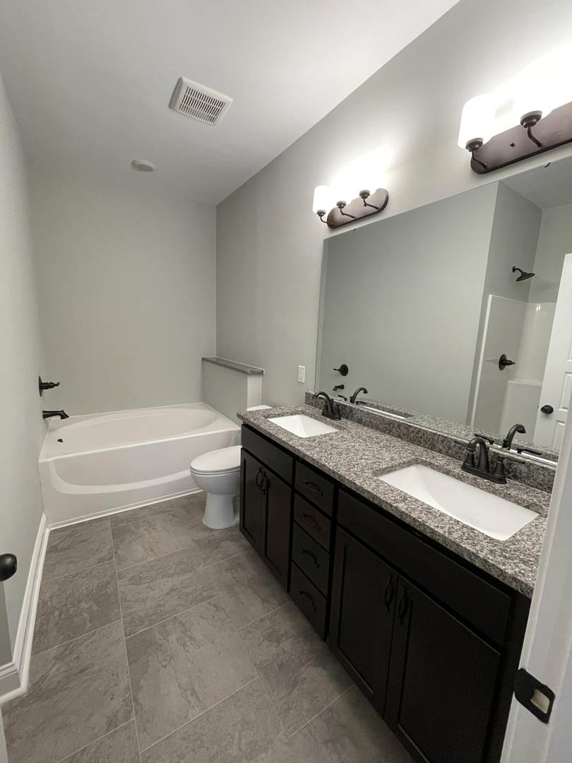 Modern bathroom featuring a white freestanding bathtub beside a toilet, rectangular sink on a stone countertop, light tile flooring, and a white wall vent