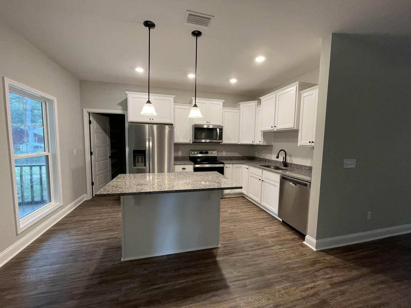 Marble kitchen island with built-in sink, stainless steel refrigerator with glass door, stainless microwave, white cabinetry, large window overlooking neighboring house, recessed