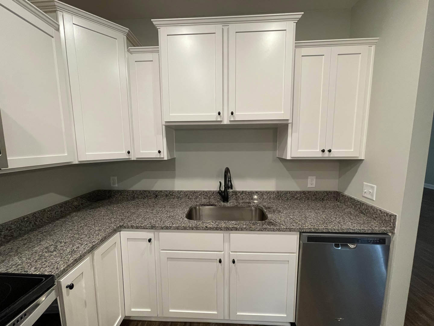 White kitchen cabinets with black handles, granite countertops, stainless steel sink, built-in dishwasher, stove top, and a white double light switch on the wall.