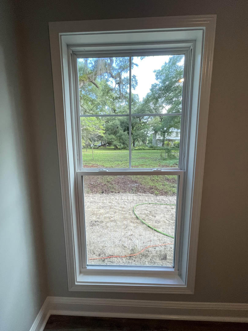 Large window with white molding overlooks green trees and a garden hose on the ground outside; interior wall and cable visible in foreground.