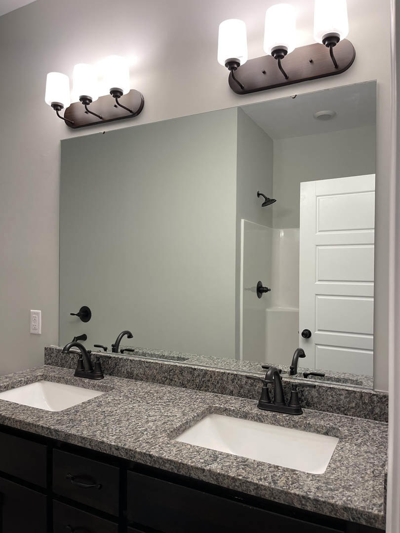 Bathroom with double sinks, wide framed mirror, quartz countertop, chrome faucets, white cabinetry, and neutral tile backsplash