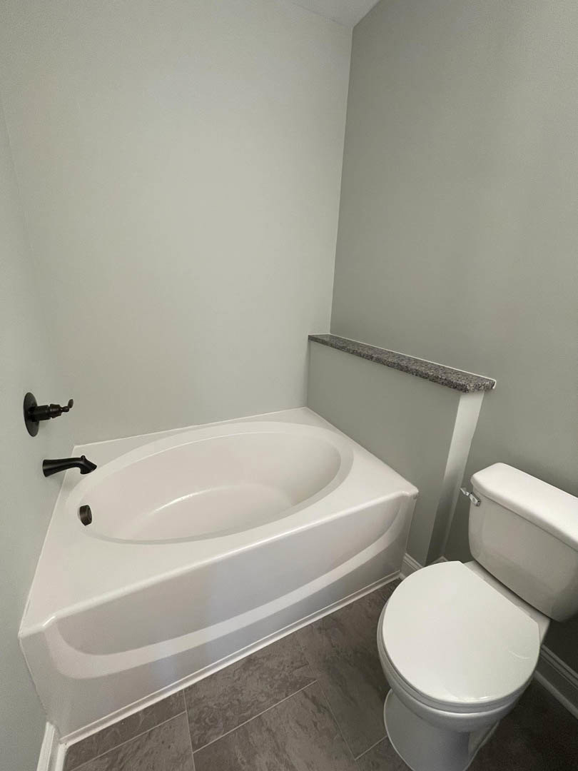 Modern bathroom featuring a freestanding white bathtub with matte black faucet, white toilet with closed lid, light-colored tile walls, and a stone countertop