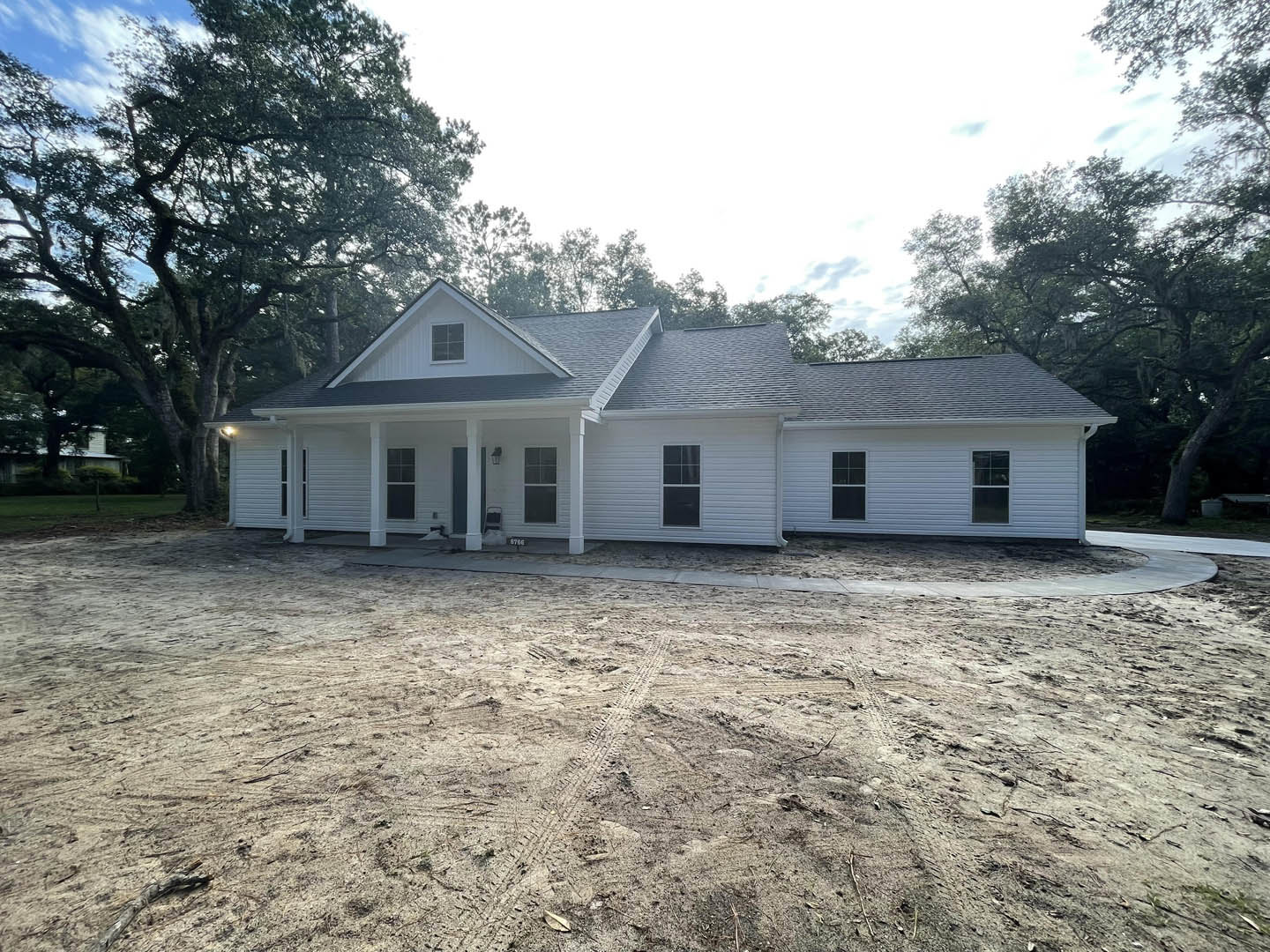 White house with black roof and white-framed window, surrounded by dirt lot with visible tire tracks, trees in background, under clear sky