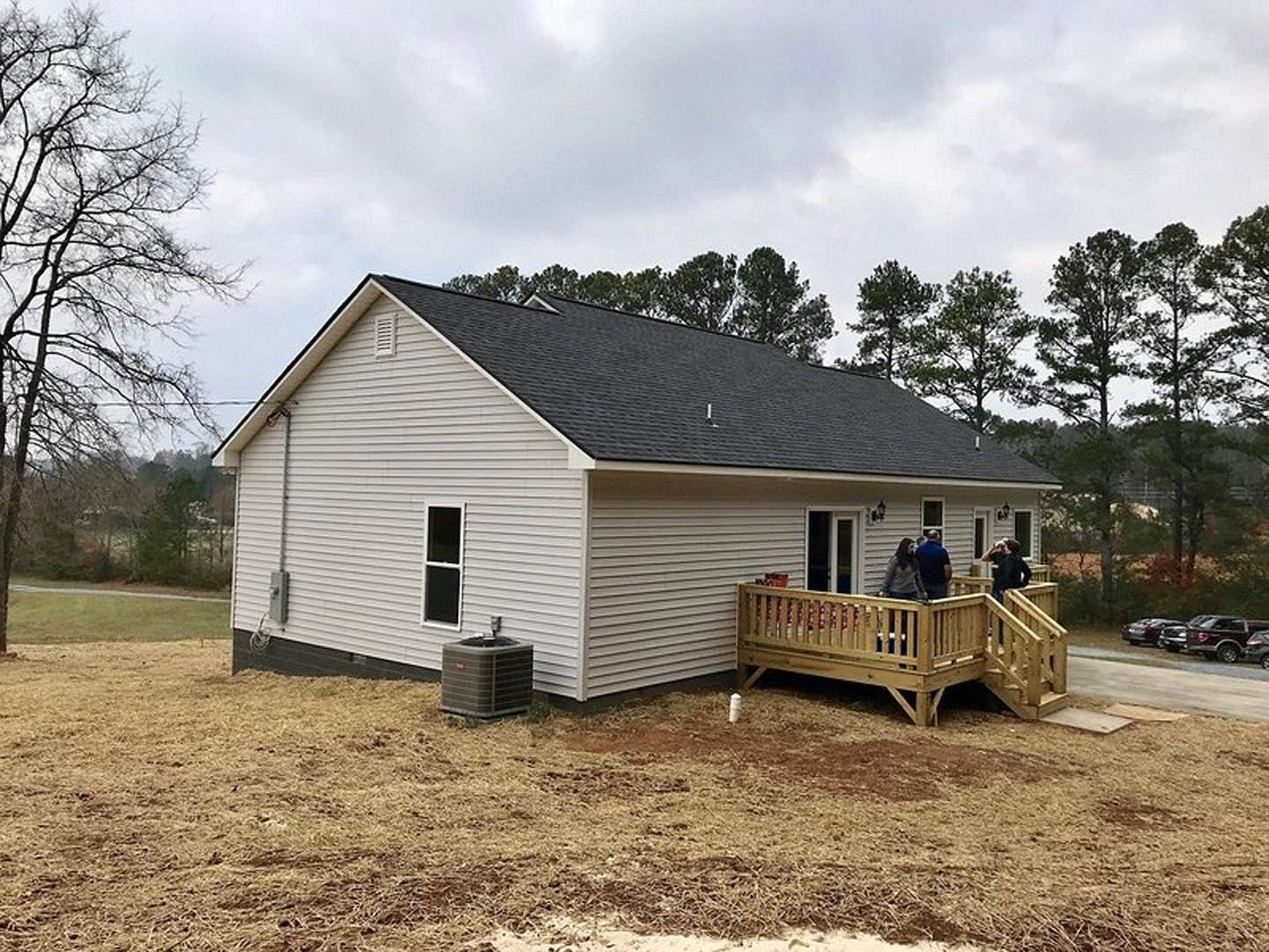 Wood deck attached to a house with group of people standing, leafless tree nearby, window and air conditioner visible on exterior wall, cloudy sky overhead.