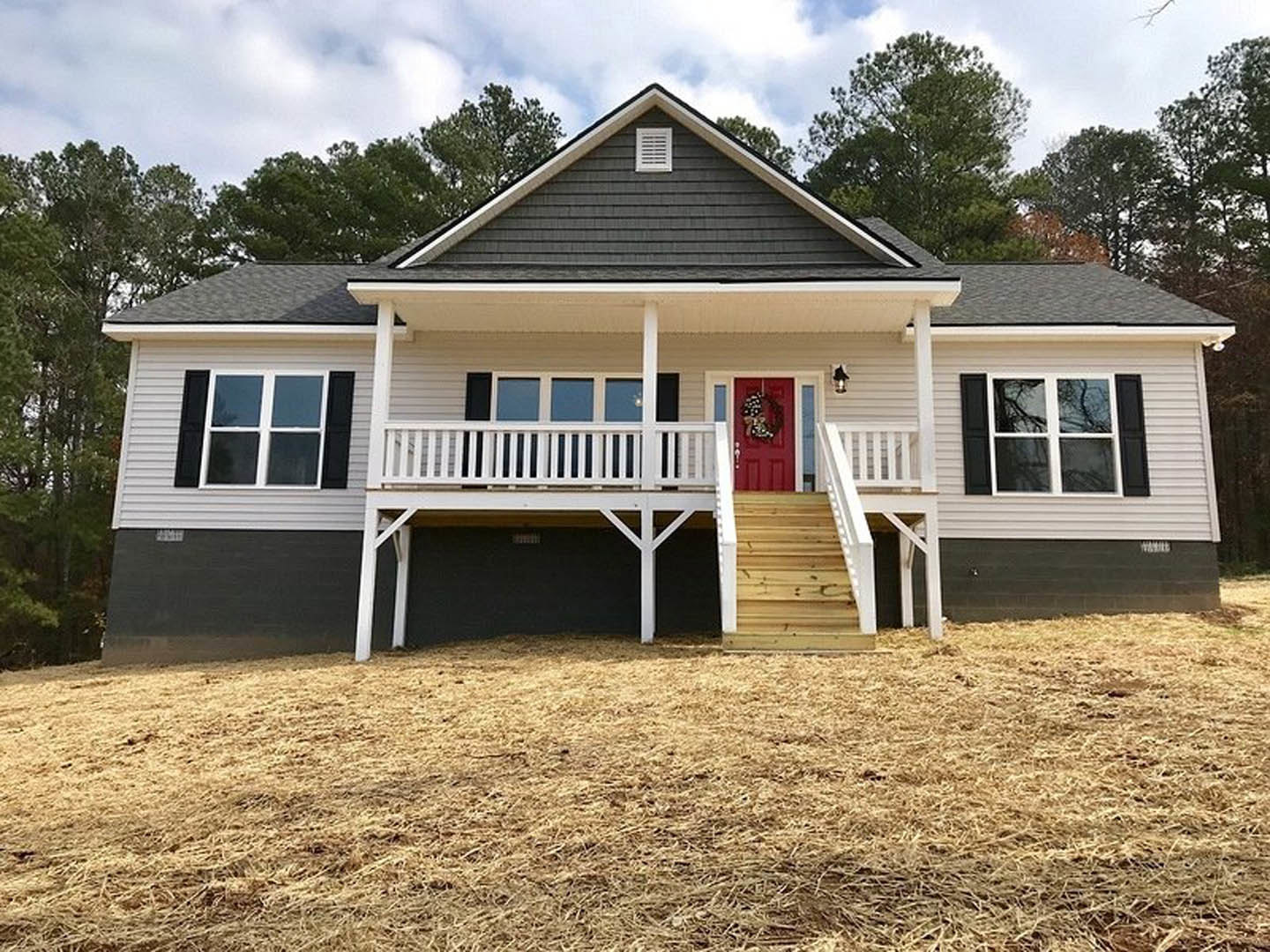 White siding house with a red front door, white framed windows, covered porch with white railings, hay scattered on the ground, leafy tree partially visible.