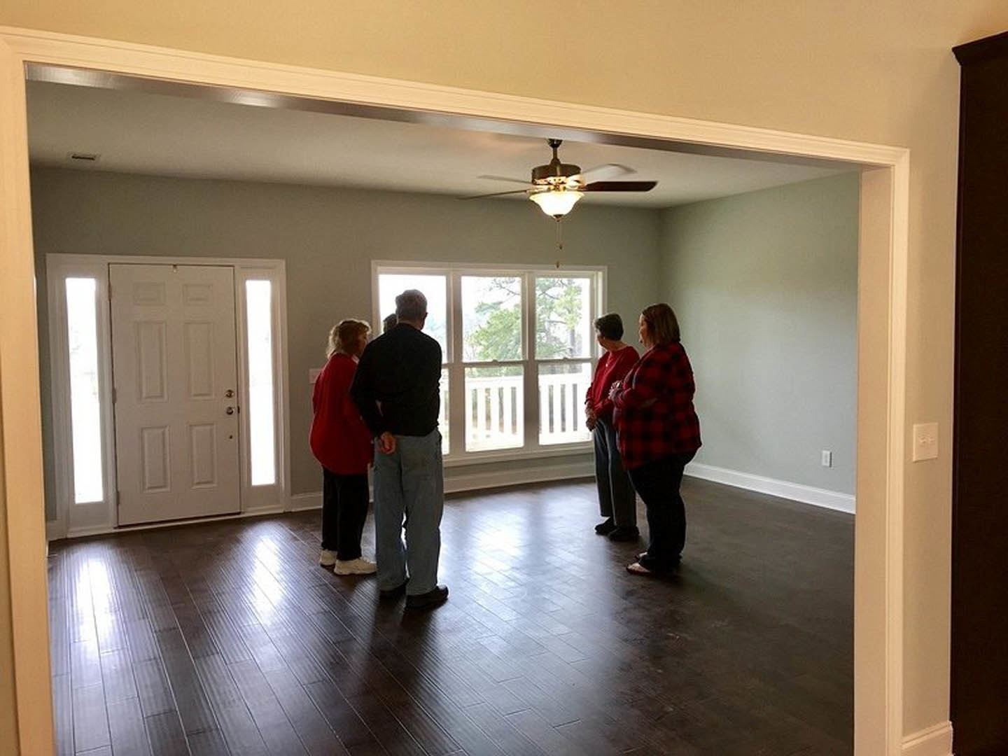 Group of people standing on light wood flooring in a room with white walls, ceiling fan with light fixture, and white door featuring two glass windows