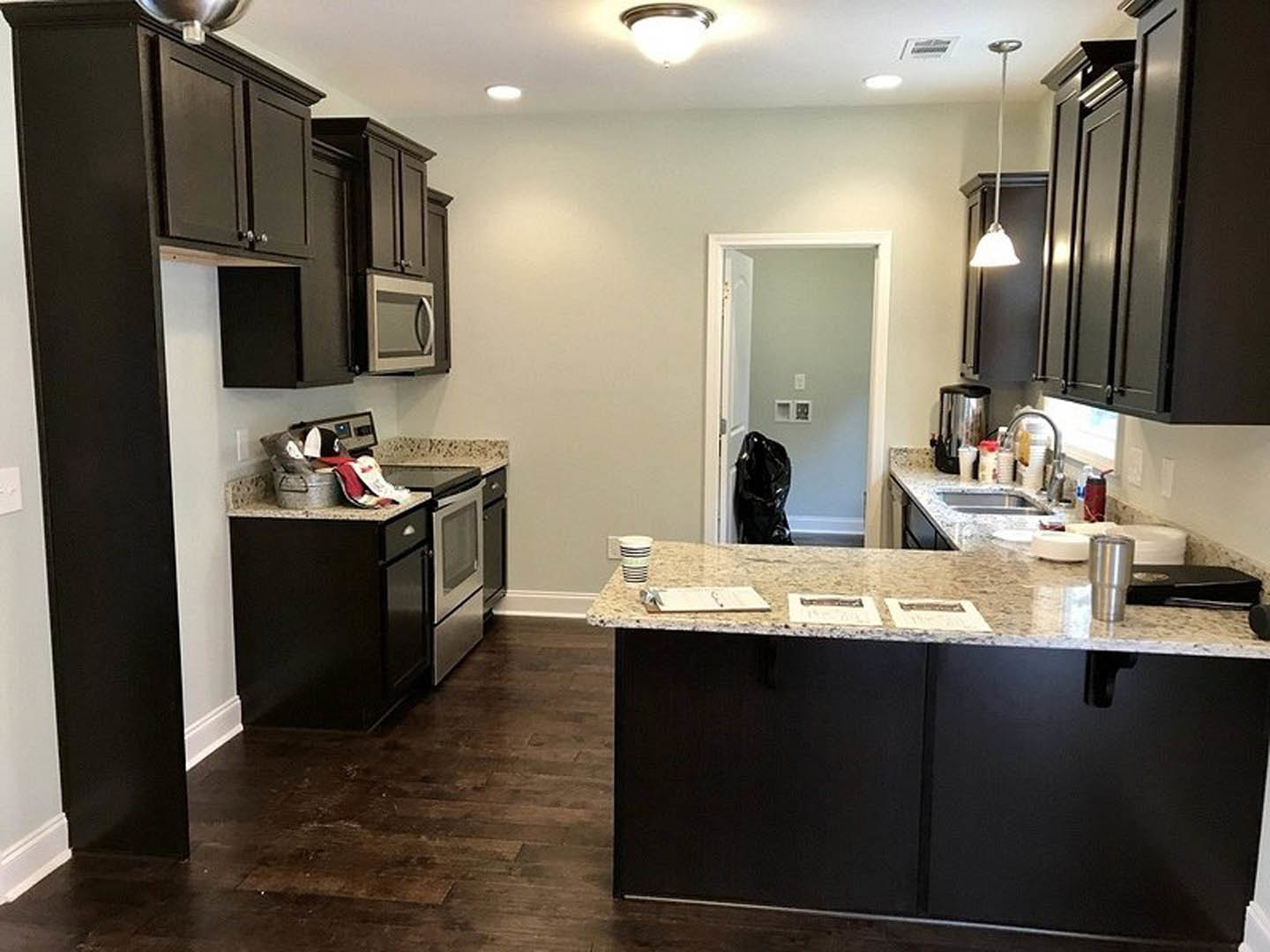 Kitchen featuring dark wood cabinets, granite countertops, stainless steel sink and faucet, built-in microwave, bell-shaped pendant light, open doorway to adjacent room, papers