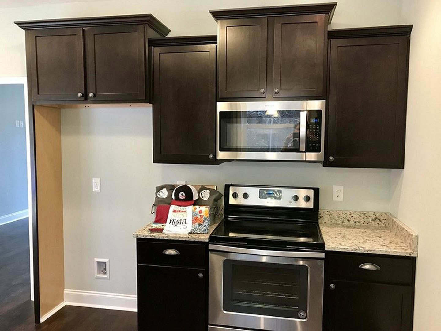 Kitchen with dark wood cabinets, stainless steel microwave above stove with open oven door, window with cracked glass, white walls, small white window, black and white hat and