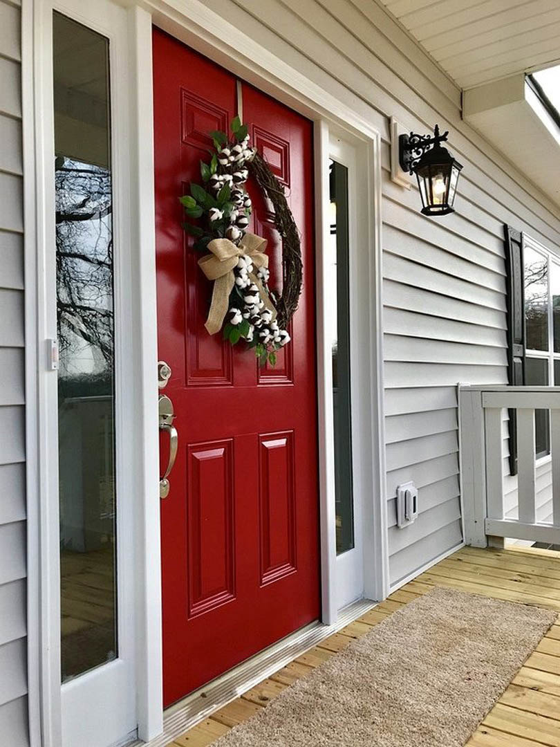 Red front door with cotton flower and burlap wreath, white siding, black door handle, outdoor entry rug, adjacent wall-mounted lamp.