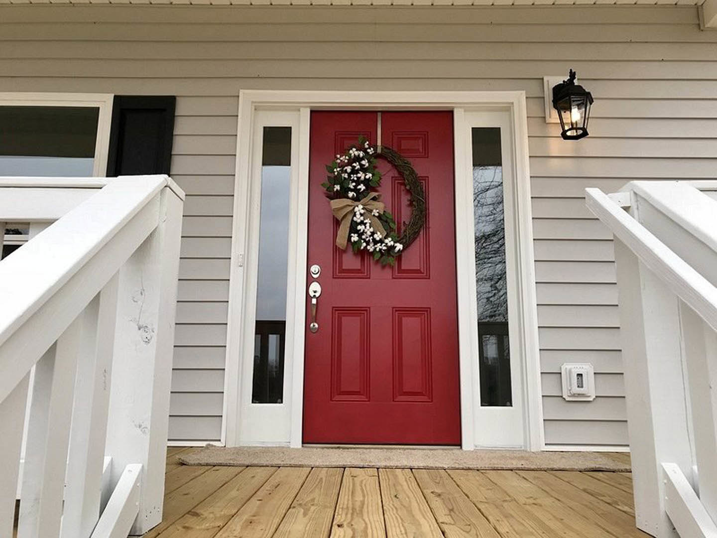 Red front door with a white floral wreath and bow, white railing and light fixture on porch, exterior entryway with stairs and door handle