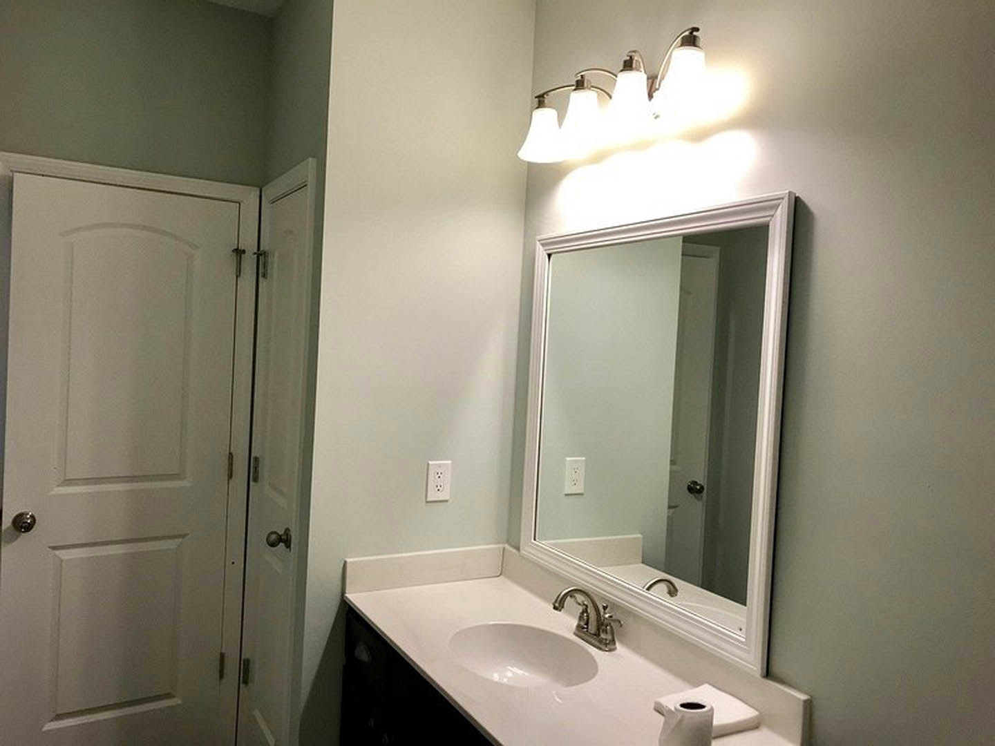 White bathroom with rectangular mirror above a marble countertop sink, chrome faucet, row of exposed light bulbs, and white door with silver knob.