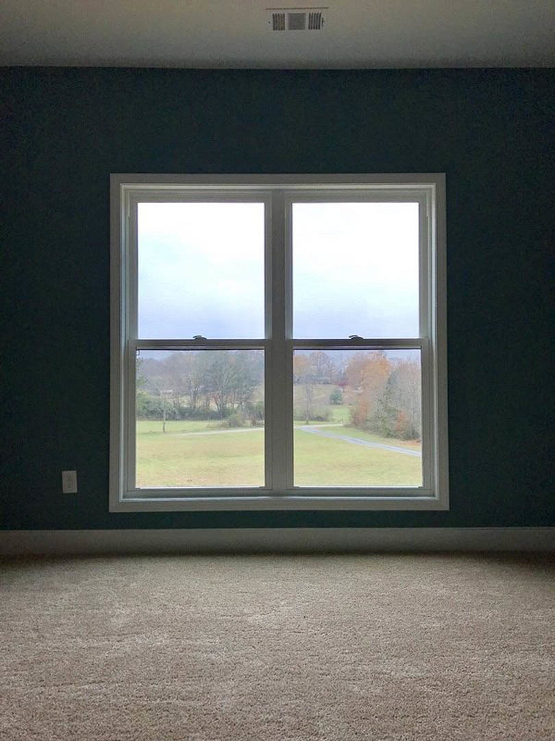 Large rectangular window set in a white wall with central vent, overlooking grassy field and trees; beige carpeted floor visible in foreground.