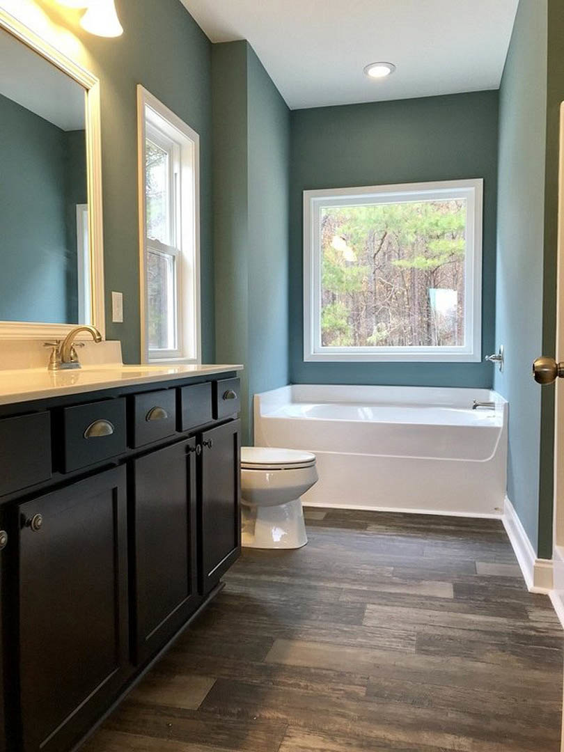 Bathroom with white bathtub beside closed toilet, wood flooring, large window revealing trees outside