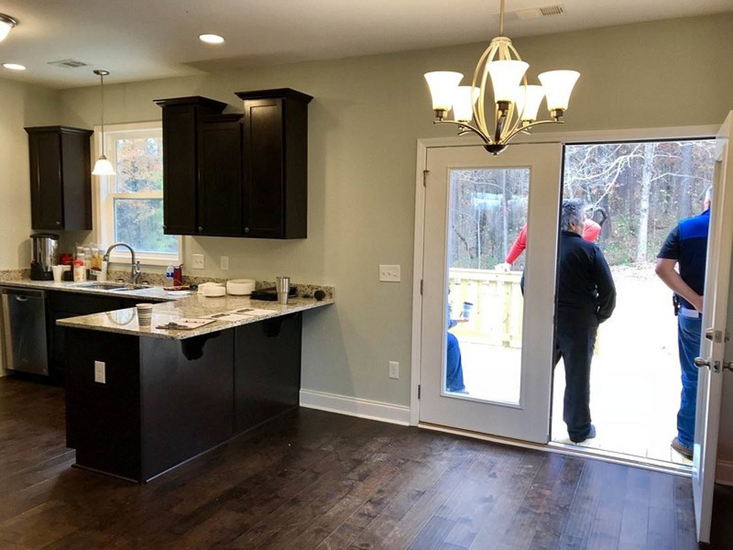Open kitchen with white cabinetry, stone countertops, stainless steel sink, and a group of people standing in the doorway; black cabinet and chandelier visible, light wood flooring