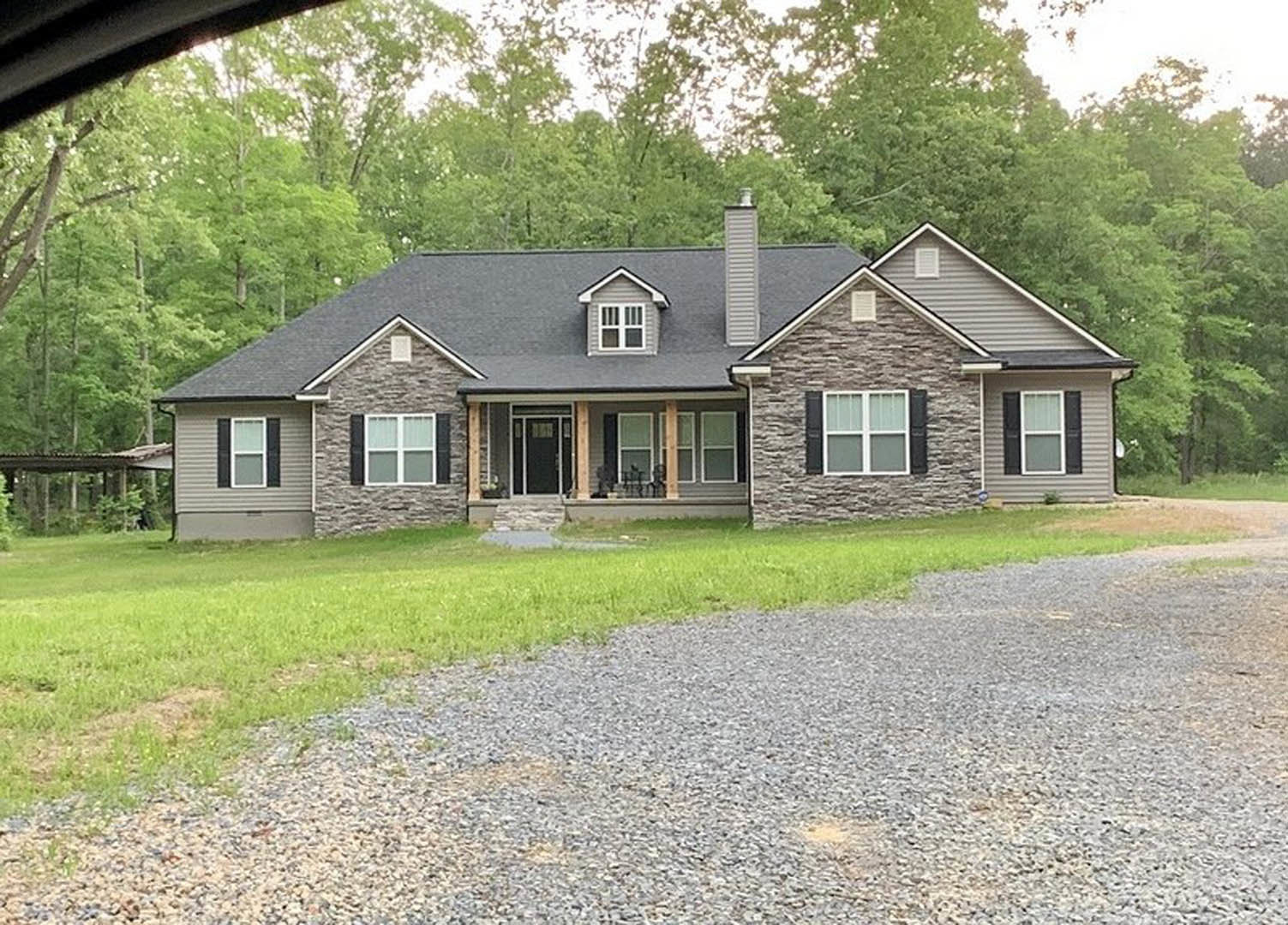 Two-story house with light siding, black-framed windows, covered front porch, gravel driveway bordered by grass, mature trees surrounding the property