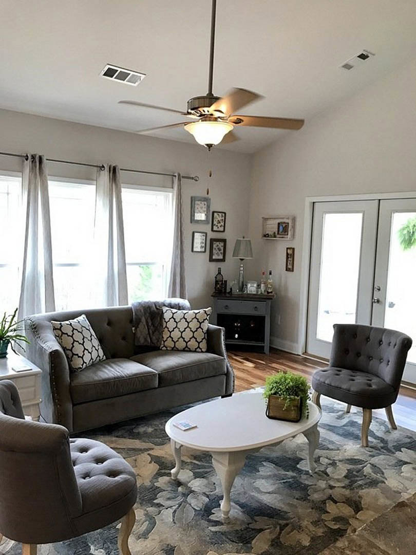Living room with light-colored couches, decorative pillows, white oval coffee table with potted plant, black accent chair, ceiling fan, and neutral walls.
