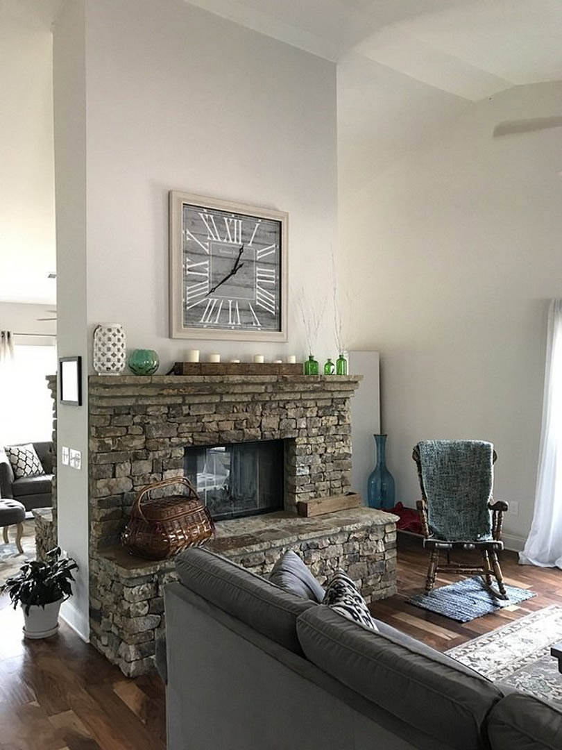 Living room with stone fireplace, gray couch with patterned pillows, wall-mounted clock, armchair draped with blanket, potted plant, woven basket on couch, hardwood flooring