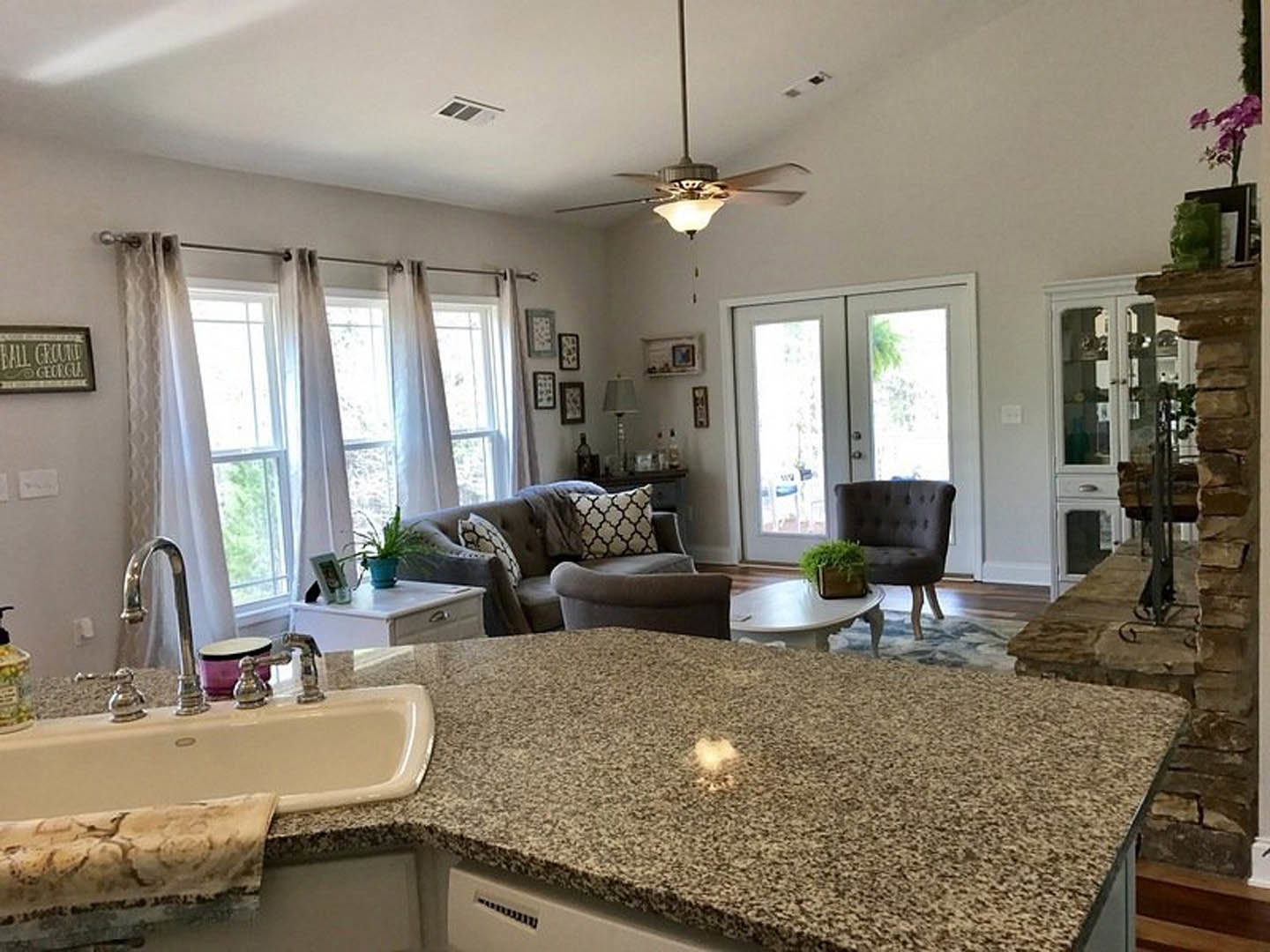 Open-concept kitchen and living room featuring white cabinetry, stainless steel sink, ceiling fan, couch with pillows, upholstered chair, double doors, and floor-length curtains