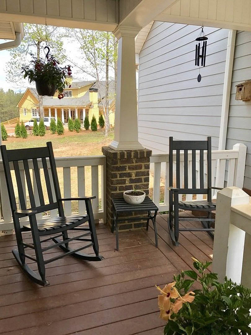 Wide front porch with black rocking chairs, white flower pots, and a small black table, surrounded by outdoor greenery.
