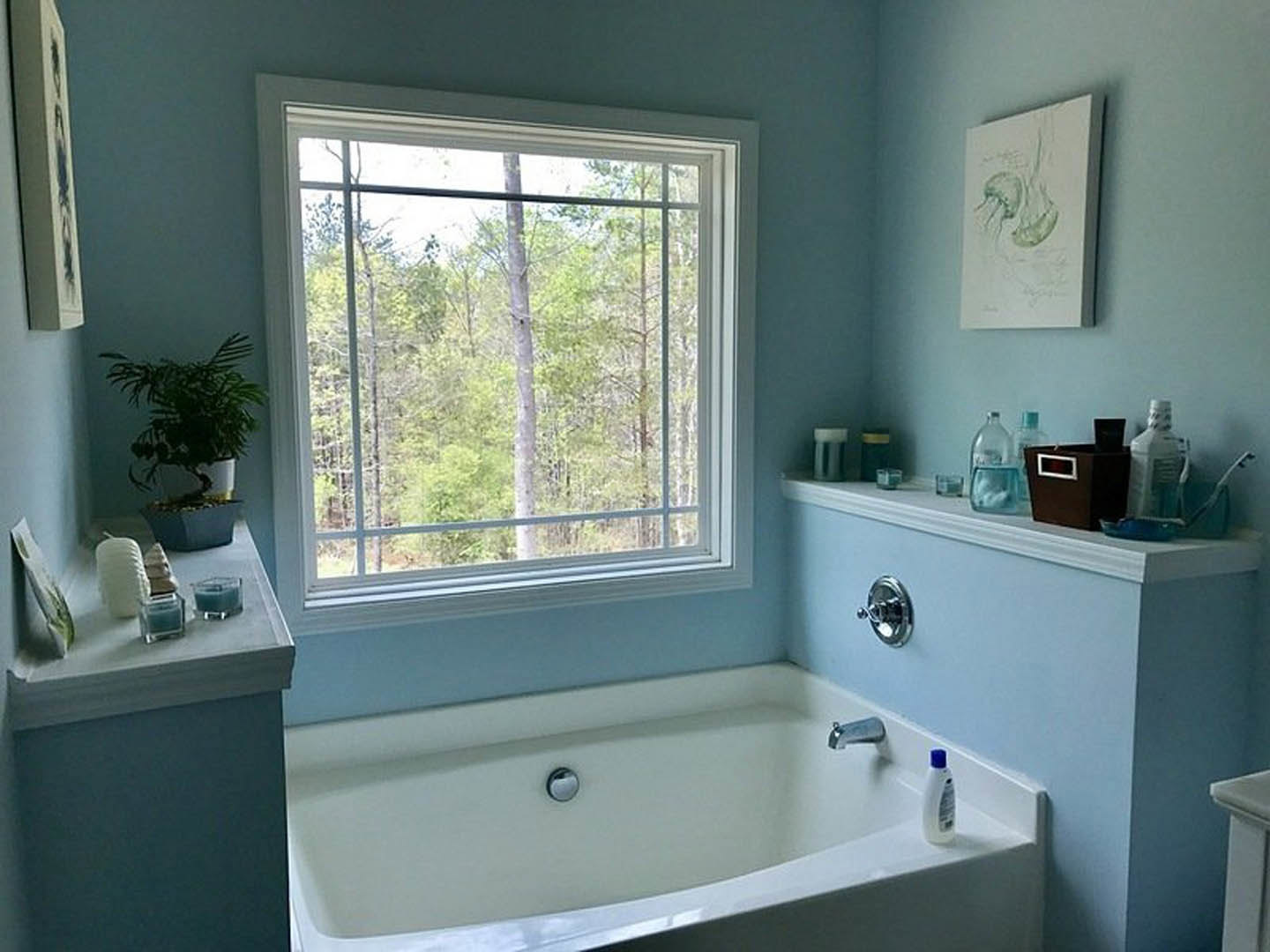 White freestanding bathtub beneath a large window with leafy trees visible outside, chrome faucet, light gray tiled walls, small potted plant on windowsill, and partial view of a