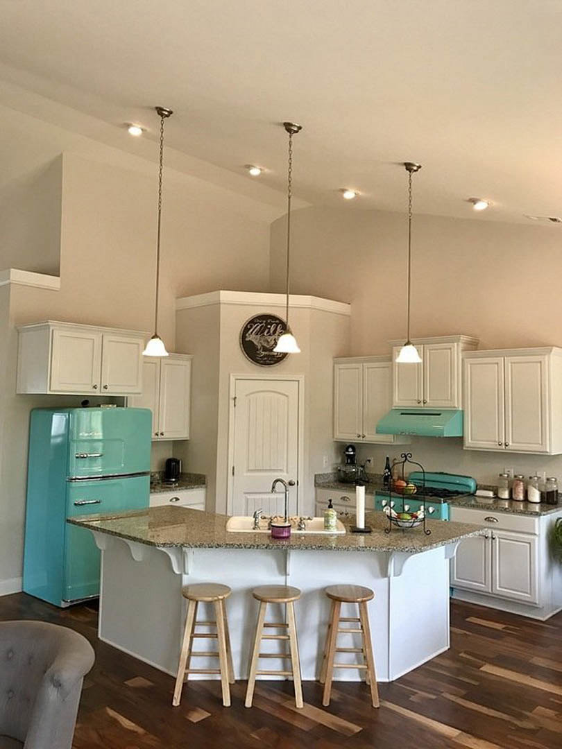 Open-concept kitchen featuring a white quartz bar counter with four modern stools, stainless steel refrigerator, white cabinetry, farmhouse sink, and a decorative dog-themed sign