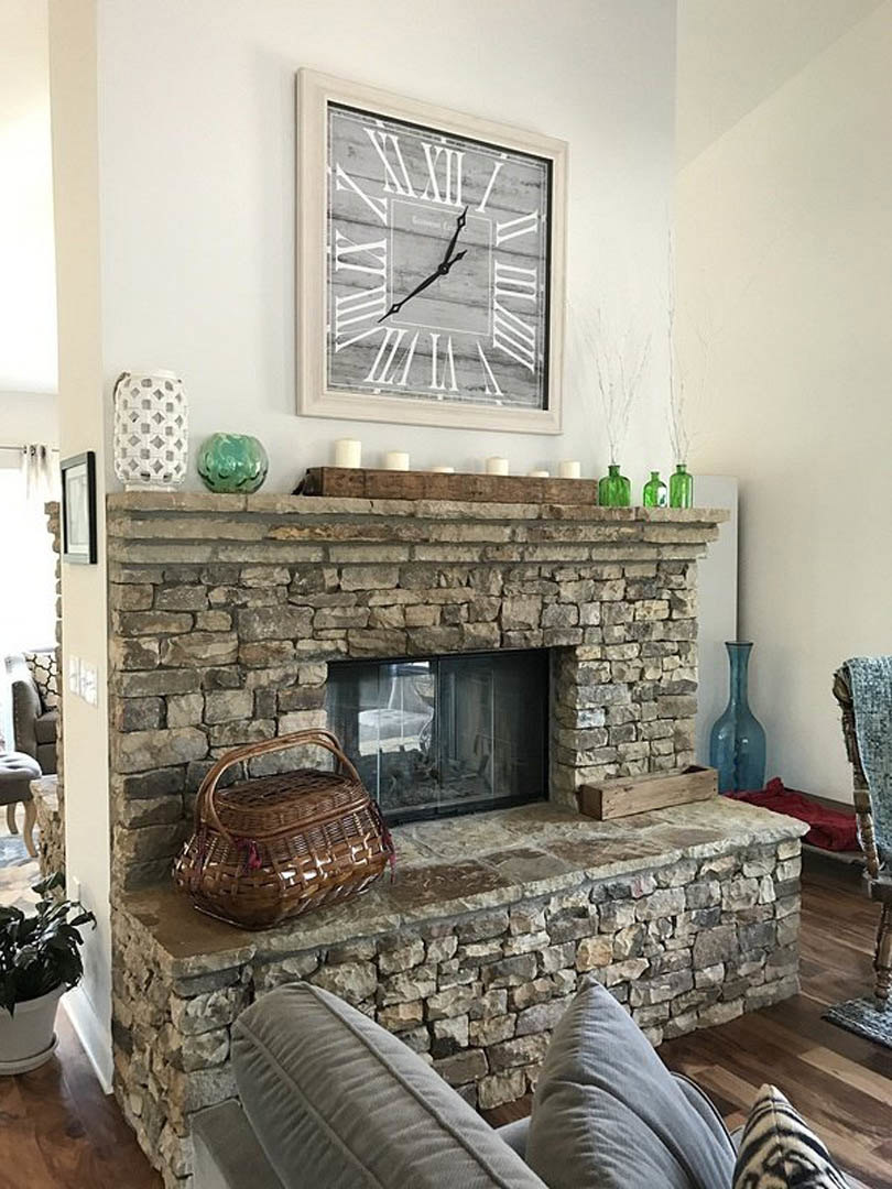 Stone fireplace with a round wall clock above, woven basket on hardwood floor, plush gray couch in foreground, potted green plant, and blue vase with white handle on mantel in cozy