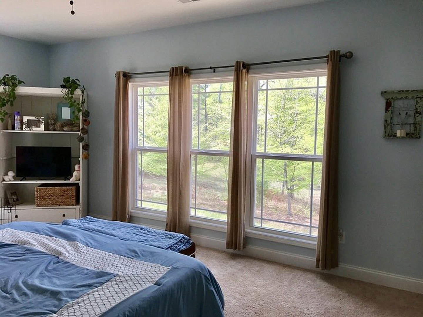Bedroom with a bed featuring a blue and white blanket, wall-mounted TV with white border, window with sheer curtains overlooking trees, and candle sconce on wall
