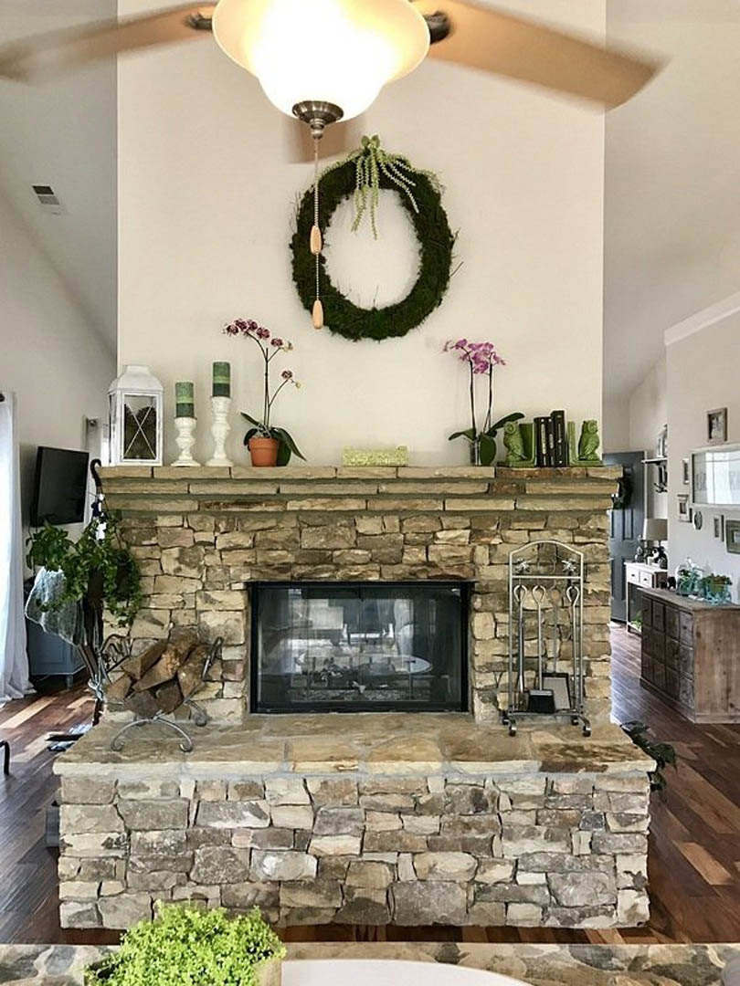 Stone fireplace with green wreath above mantel, wood hearth, nearby window with table and chairs, potted plant and fireplace tools on floor