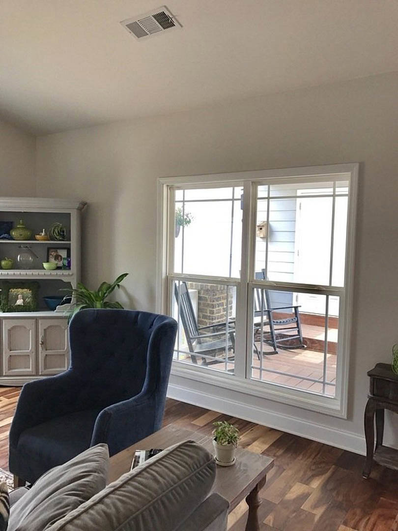 Living room with wood flooring, blue accent chair beside a small table, neutral couch, potted plant, exposed brick wall, large window with outdoor rocking chair visible