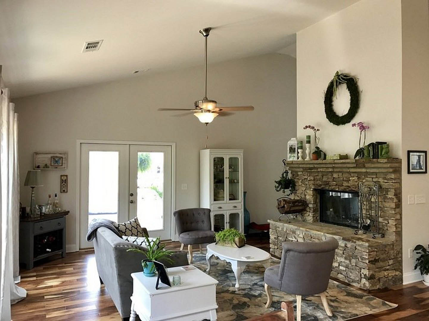 Living room with laminate flooring, white door with glass windows, grey chair, white table holding a black tablet, fireplace with white mantel, ceiling fan with light fixture, and