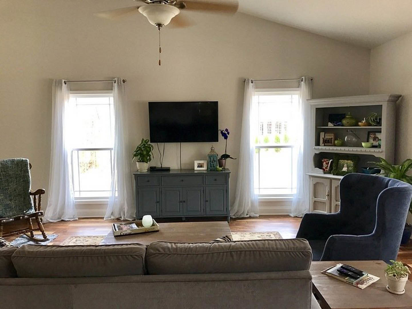 Living room with light-colored couch and two pillows, blue accent chair, wall-mounted TV with white border, window with sheer white curtains, ceiling fan, hardwood floor