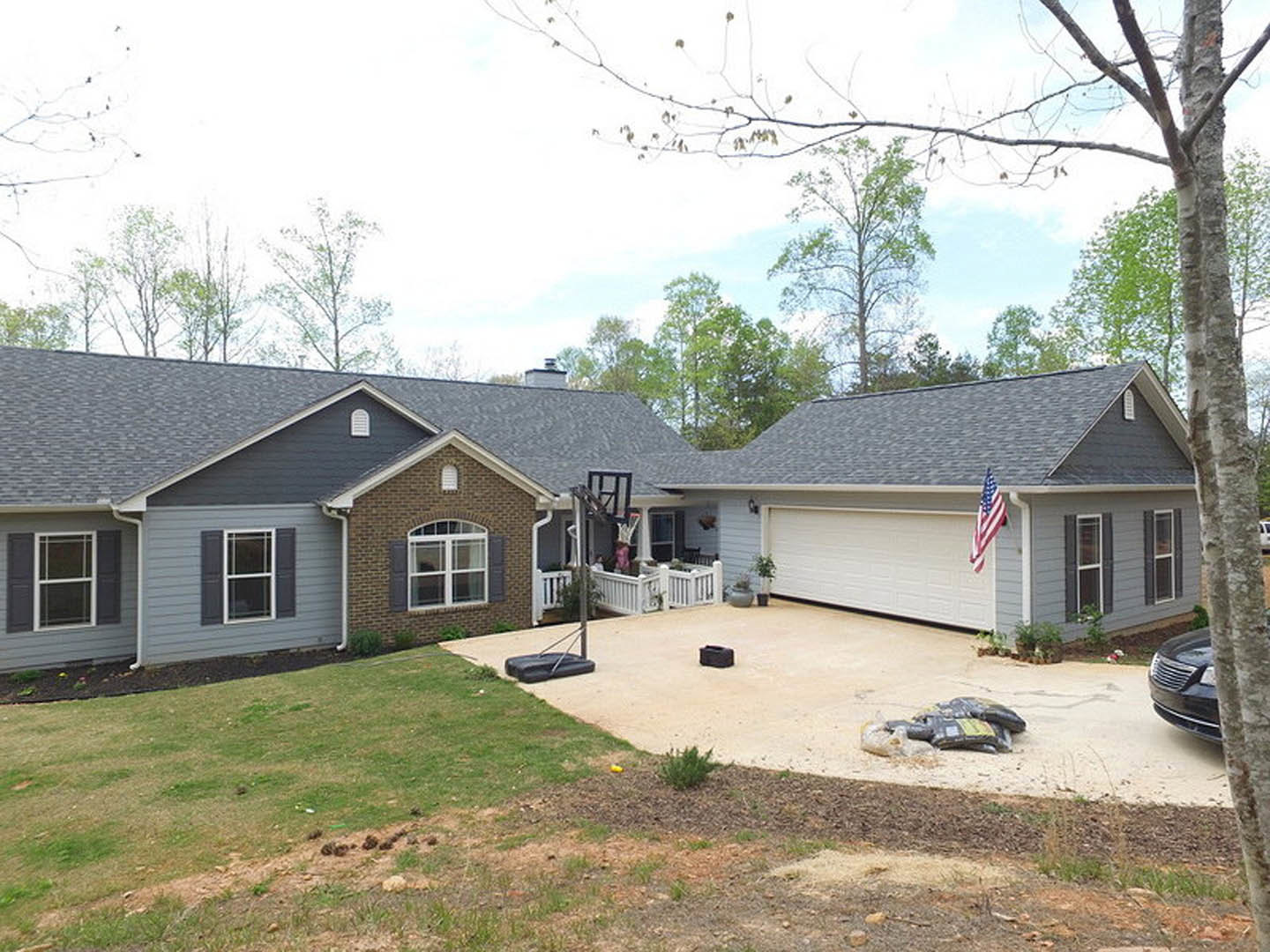 Two-story house with attached garage, manicured lawn, basketball hoop above driveway, white-framed windows, green bushes along foundation, American flag mounted near entrance.