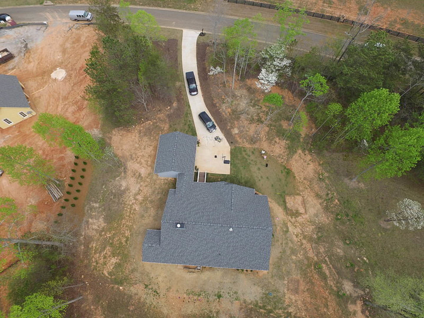 Modern two-story home with gray shingle roof, concrete driveway, parked cars, manicured lawn, and mature trees bordering the property