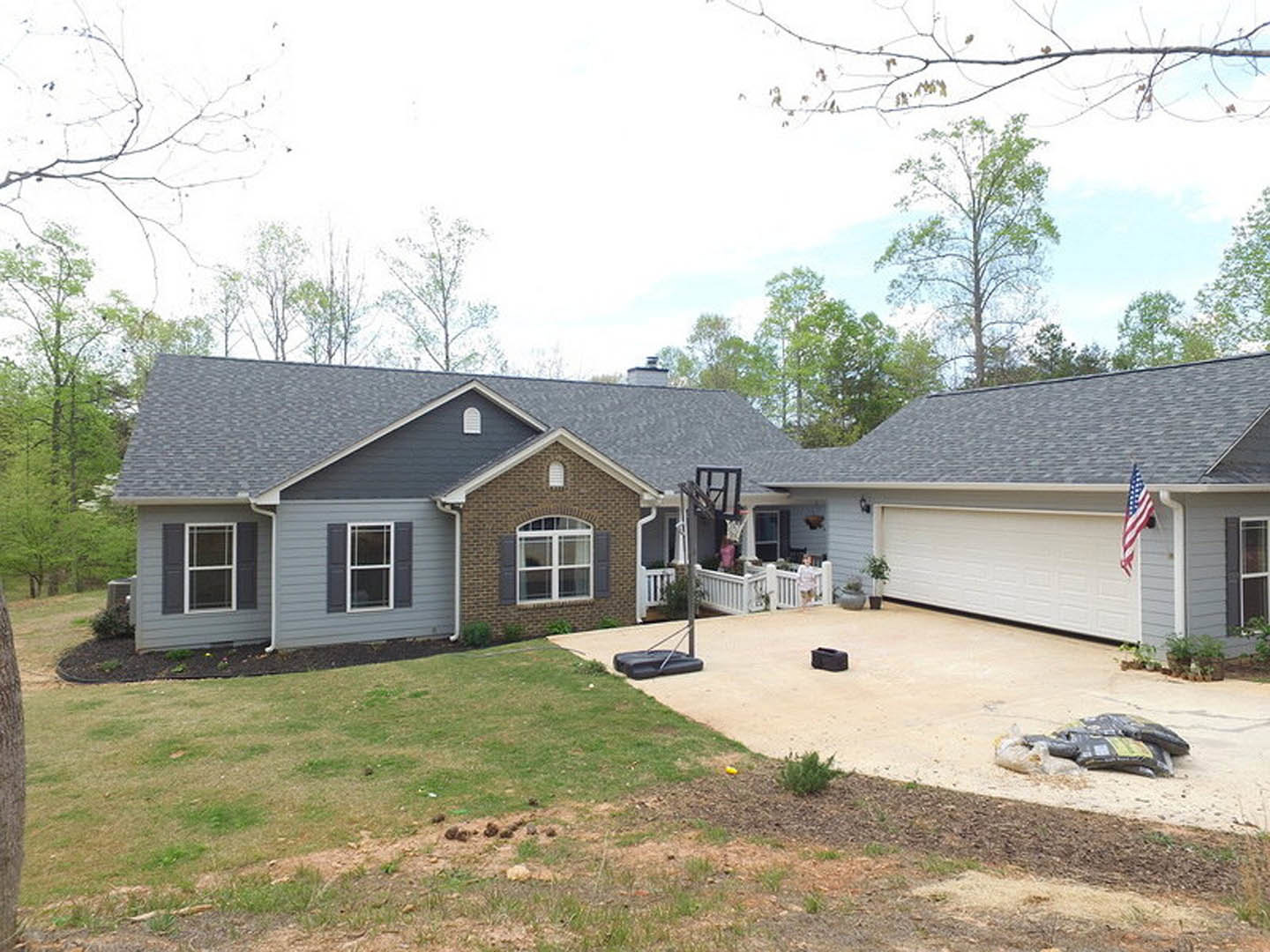 Two-story house with white-framed windows, gray roof, concrete driveway, green bush planted in mulch, basketball hoop near garage, blue sky overhead
