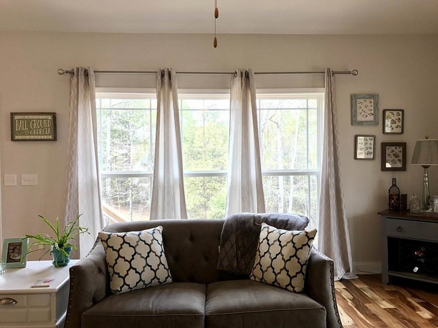 Light gray couch with patterned pillows positioned in front of a large window with sheer curtains, potted plant beside the seating area, silver floor lamp with slender stem
