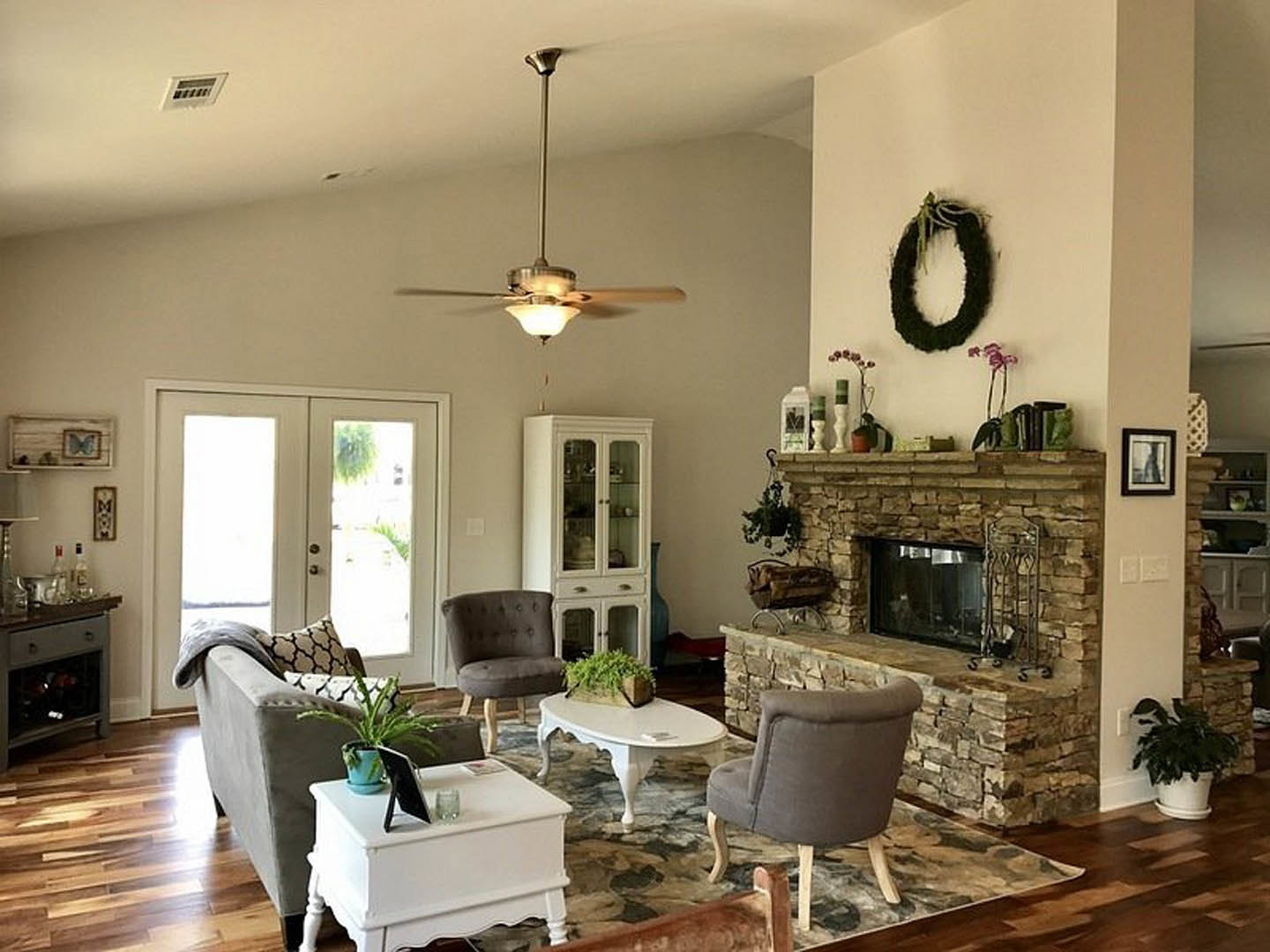 Living room with stone fireplace, gray couch, white table holding black tablet, ceiling fan with light fixture, framed portrait on wall, wall vent visible