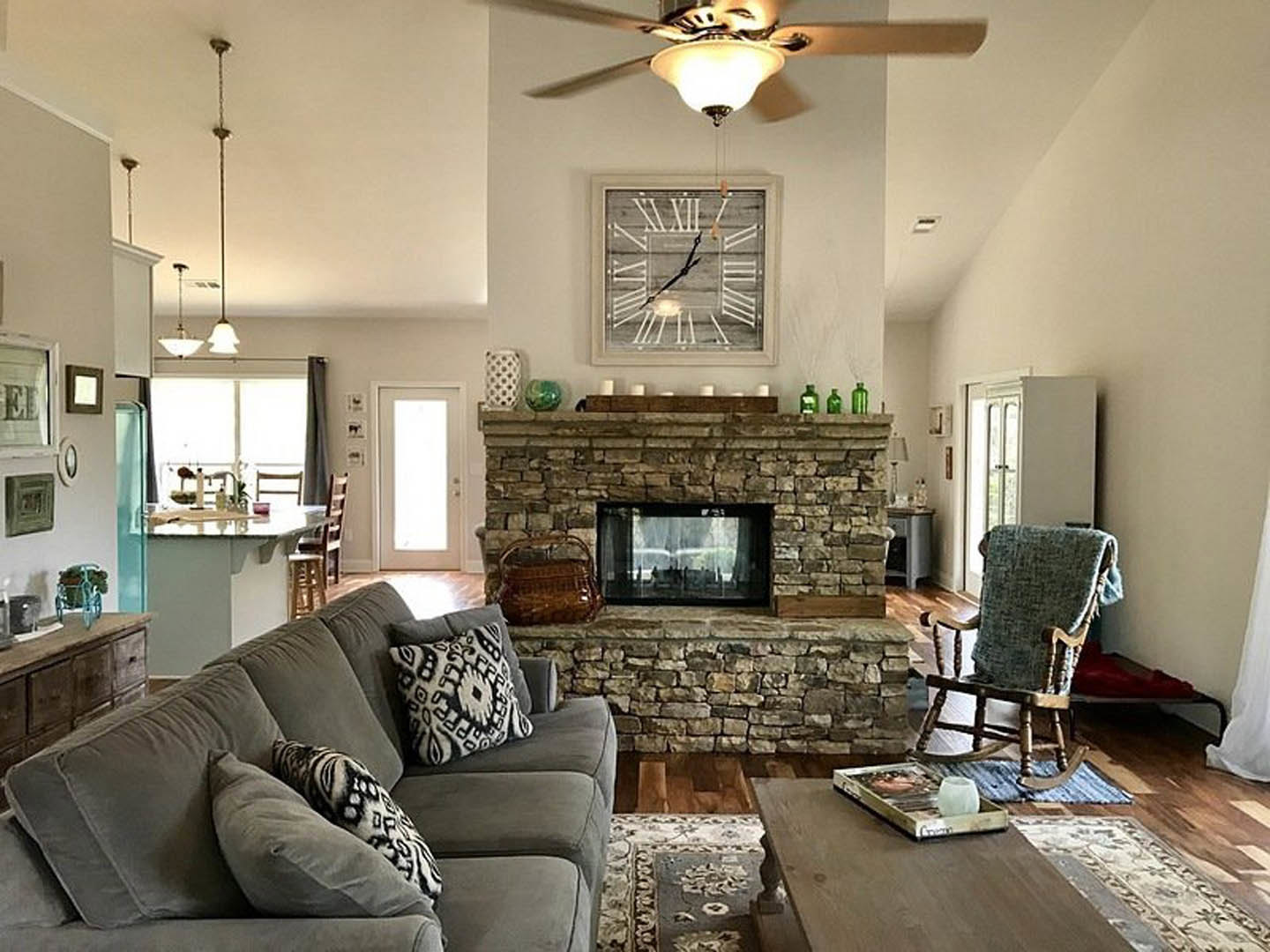 Living room with stone fireplace, beige couch with patterned pillows, wooden rocking chair, wall clock with roman numerals, hardwood floors, and large window with sheer curtains