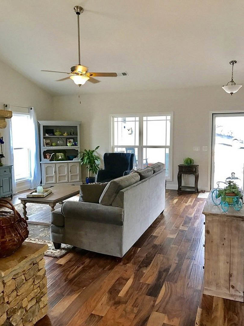 Living room with gray couch, wood laminate flooring, ceiling fan with light, green potted plant, woven basket, and decorative bicycle planter against neutral walls