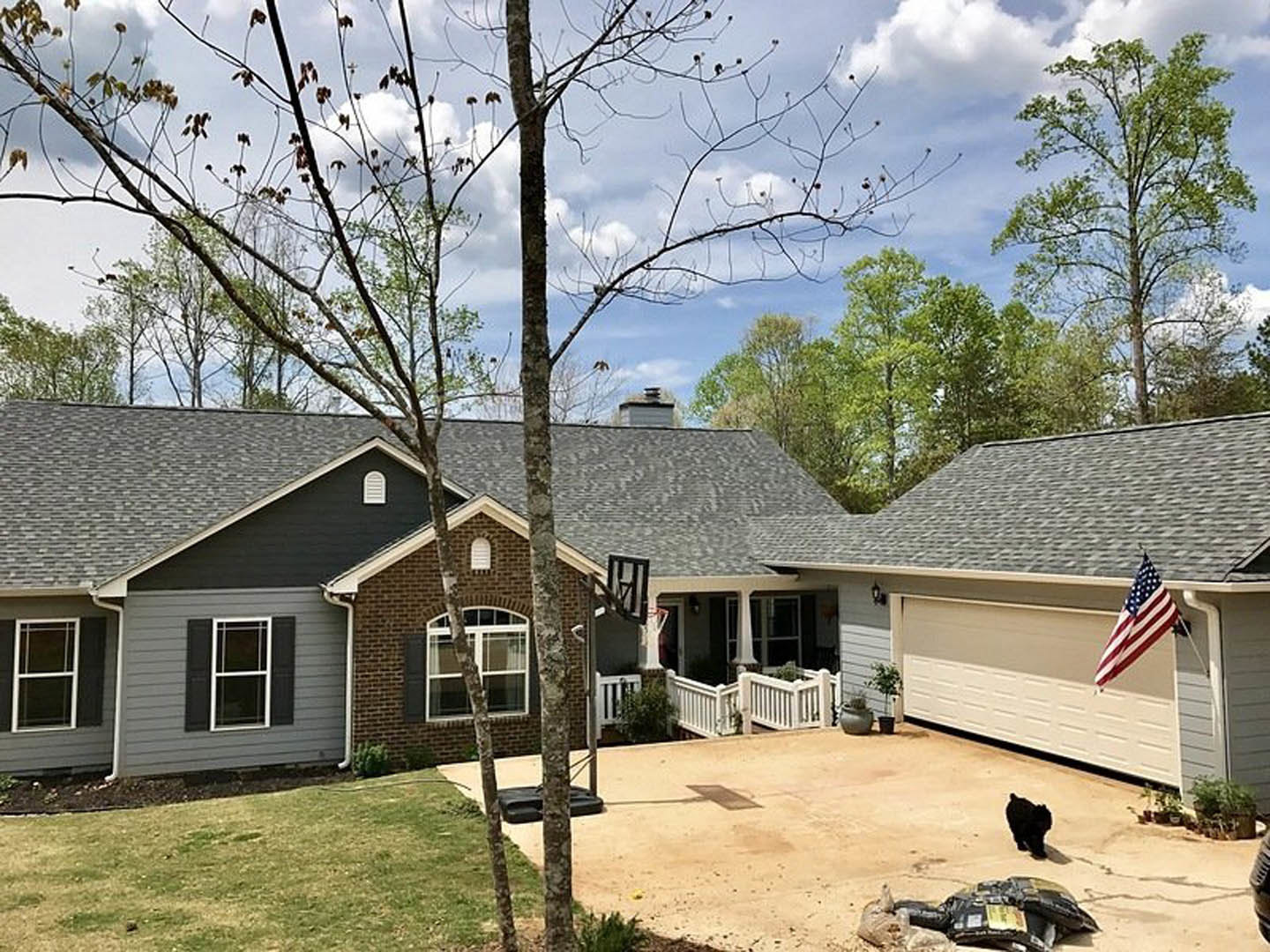 Two-story home with gray siding, attached garage, American flag mounted near entry, white-framed windows, gable roof, and landscaped front yard with trees under partly cloudy sky