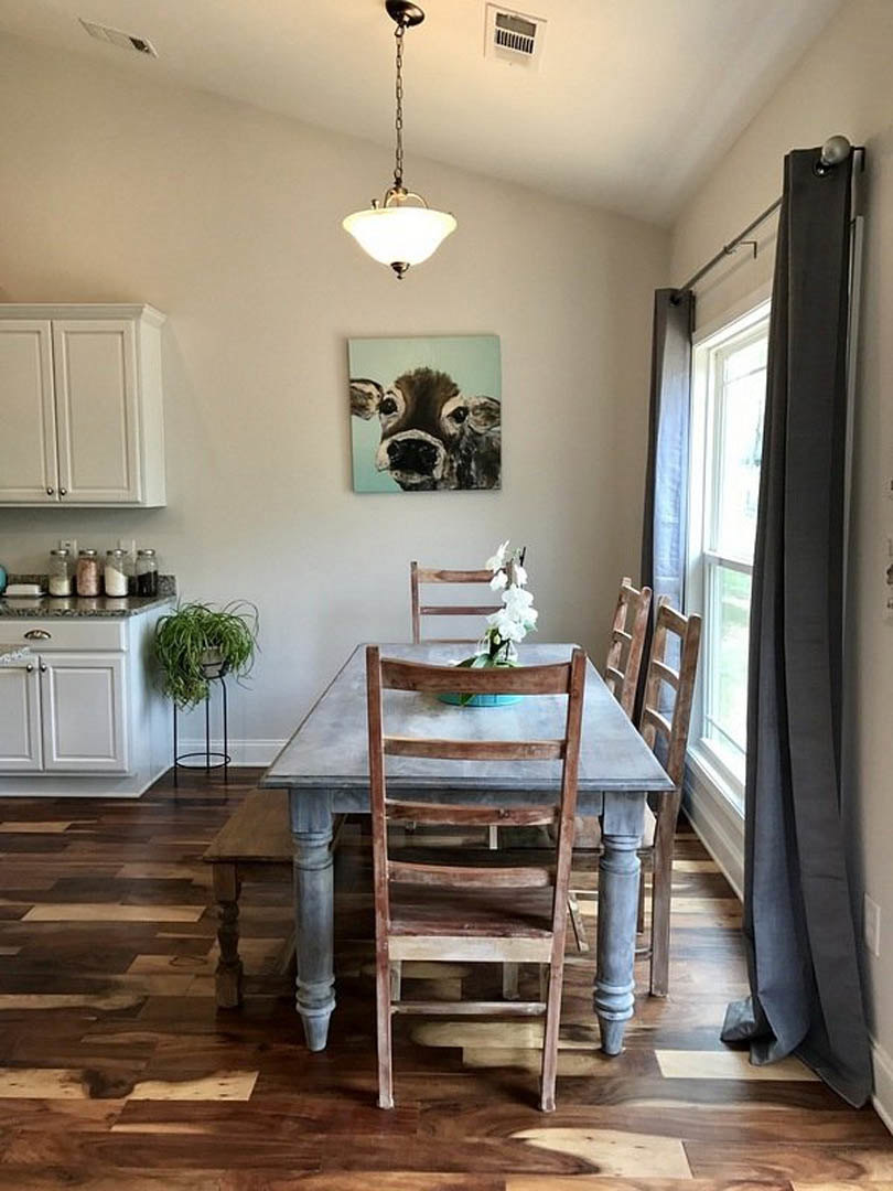 Wood dining table with matching chairs set on hardwood floor in open kitchen, white cabinetry, pendant light fixture overhead, potted plant near window with sheer curtain.