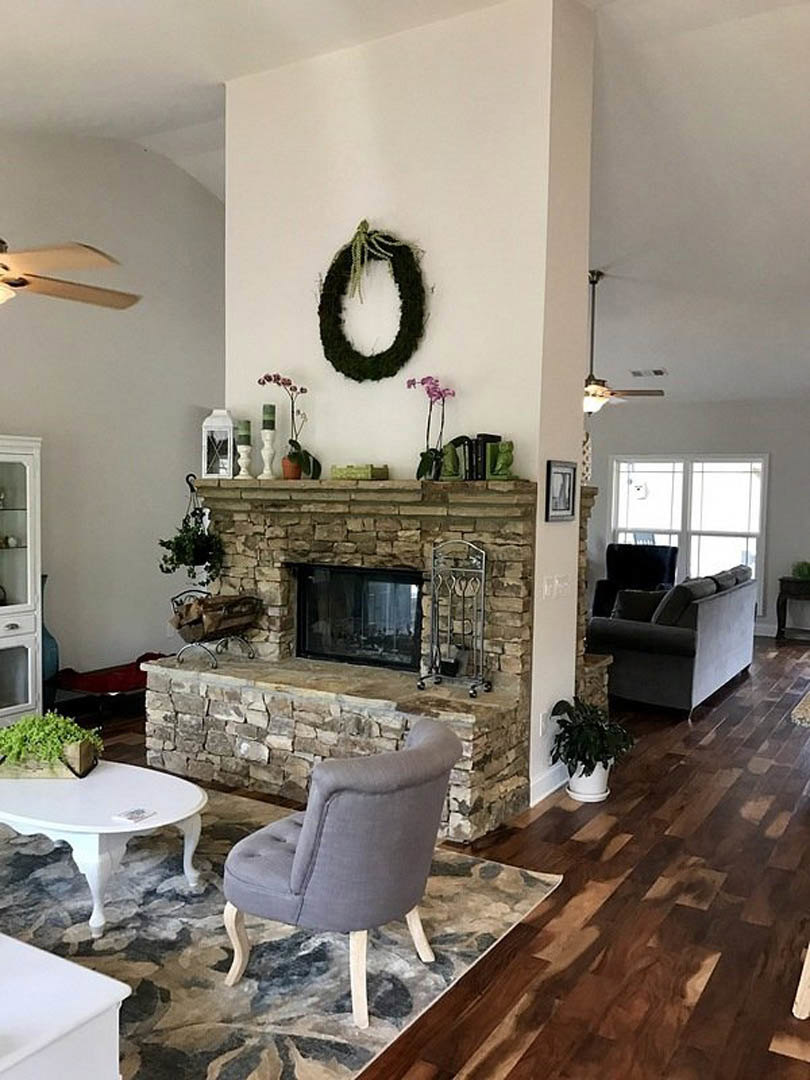 Living room with light-colored couch, grey accent chair, white coffee table, fireplace with black wreath and green plant above, potted plant in white pot, hardwood floors, neutral