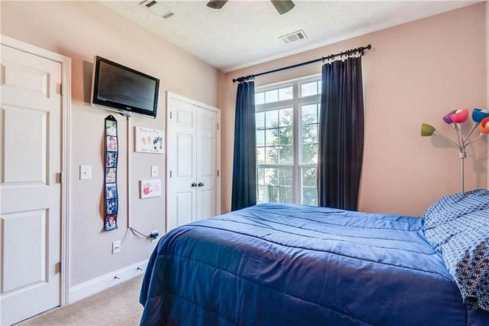 Bedroom with blue bedspread, wall-mounted television, window with blue curtains, black curtain, white door, and neutral walls