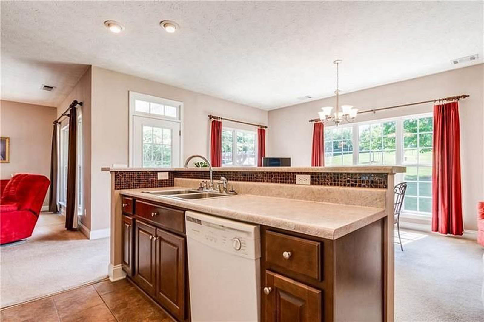 Spacious kitchen featuring a large central island with white countertop, adjacent dining area with red chairs, cabinetry in light wood finish, stainless steel dishwasher, and