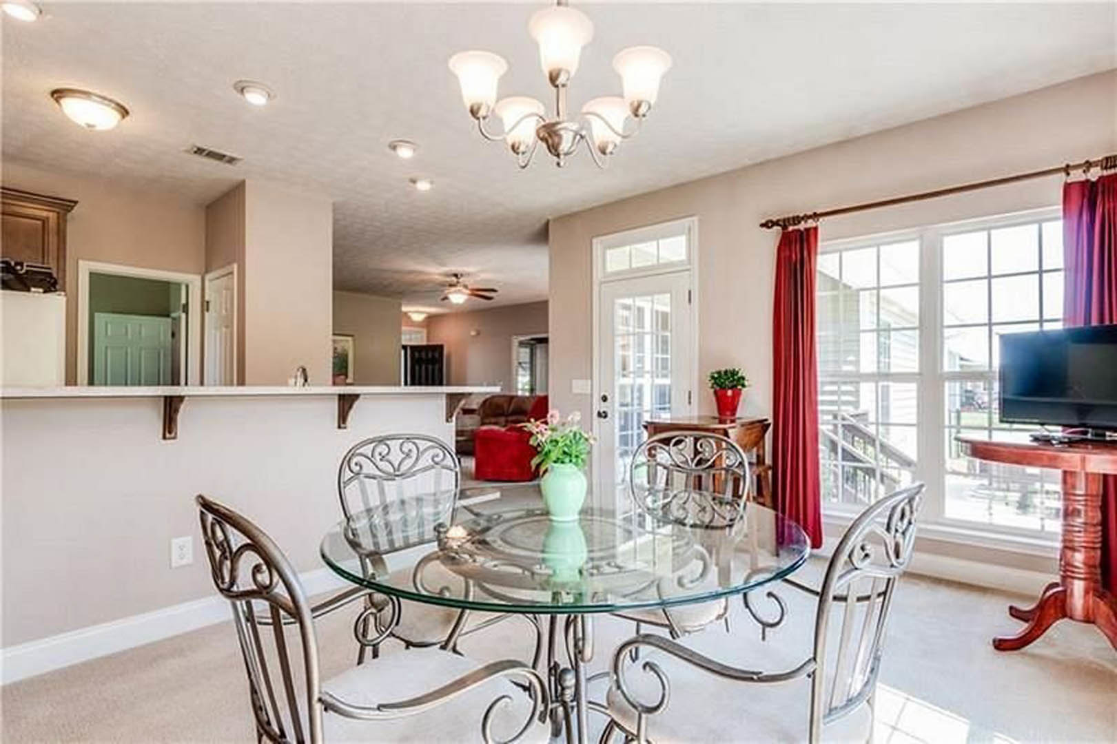 Glass dining table with upholstered chairs, decorative vase centerpiece, modern chandelier overhead, neutral walls, wood flooring, and built-in cabinetry in background.